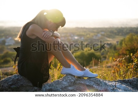 Pretty sad woman in black short summer dress sitting on a rock thinking outdoors at sunset. Fashionable female contemplating in warm evening in nature.