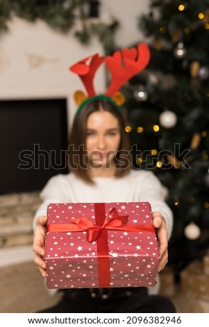 The young girl gives a red Christmas gift on a camera. Happy smiling woman with deer antlers on head sit near a festive tree