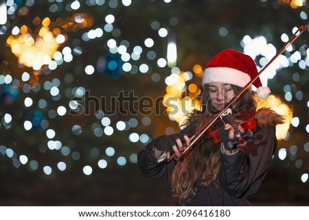 girl playing the violin in front of a Christmas tree. The lights on the Christmas tree are blurred and form a pleasant background. The girl is wearing a Christmas hat.