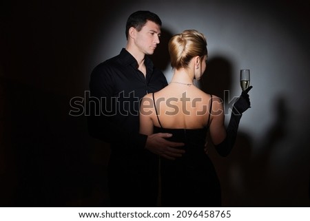 An attractive couple in black. Stylish and elegant young man and woman in the spotlight. Beautiful blonde in a long dress with a glass in her hands
