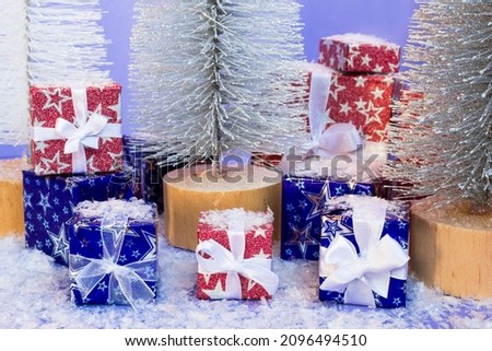Gift boxes wrapped in shiny blue and red paper with artificial snow and Christmas trees on purple background.