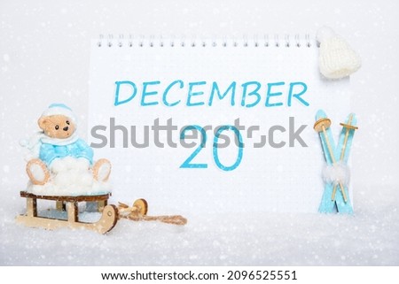 December 20th. Teddy bear sitting on a sled, blue skis and a calendar date on white snow. Day 20 of month.
