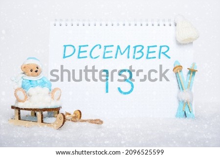 December 13th. Teddy bear sitting on a sled, blue skis and a calendar date on white snow. Day 13 of month.