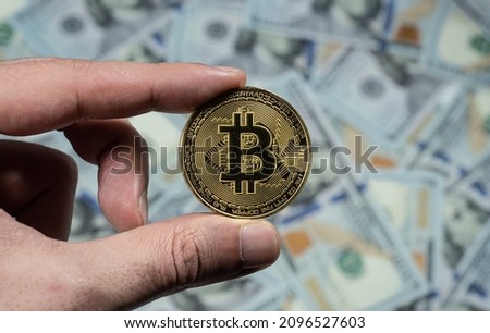 Male hand holds gold bitcoin coin, one hundred dollar bills background the gold bitcoin coin.