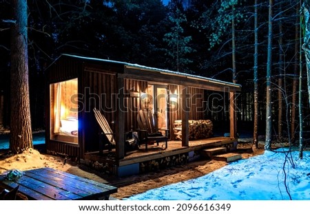 Wooden cottage at night in forest in winter. White snow around wooden house at night. Light goes through window and lies on snow