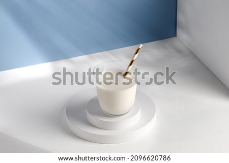 Milk in glass with a straw on white podiums. Natural milk on pedestal in sunlight. Shadow on blue wall.
