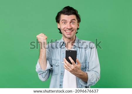 Joyful young brunet man 20s wearing casual clothes white t-shirt denim shirt posing using mobile cell phone typing sms message doing winner gesture isolated on green color background studio portrait