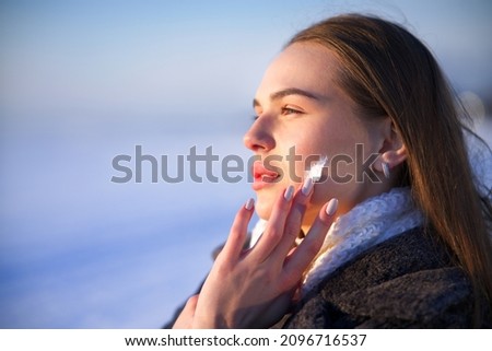 Portrait of beautiful woman applying cosmetic skin cream on face and hands, protecting from cold weather at winter season. Pretty girl take care of her skin outdoors on snowy nature background. 
