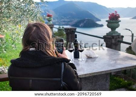 Pretty female tourist taking pictures with her phone of Lake Como (Italian Alps) while drinking a glass of wine at vacation home.