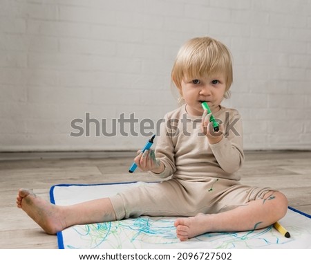 A little cheerful girl in pajamas is sitting on the floor and gnawing on a water marker. The face and clothes are stained with paint