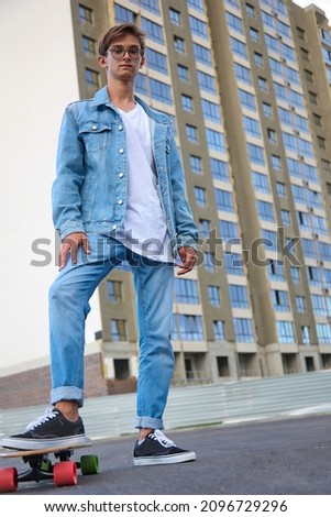 Modern teenager in blue jeans and sneakers stands with one foot on a skateboard. A multi-storey building is depicted in the background.