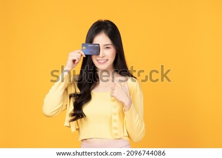 Cheerful young asian woman in yellow T-shirt holding bank card isolated on yellow background. Asian girl point to credit card.
