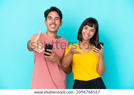 Young mixed race couple holding mobile phone isolated on blue background shaking hands for closing a good deal