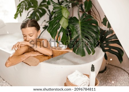 Portrait of playful young lady smiling aside while leaning on side of white bath decorated with tropical plant. Wellness, beauty and care concept