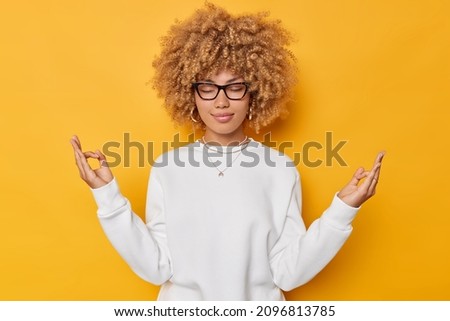 Portrait of beautiful woman with curly hair practices yoga indoors holds hands in mudra gesture keeps eyes closed fingers together wears white jumper isolated over yellow background breathes deeply