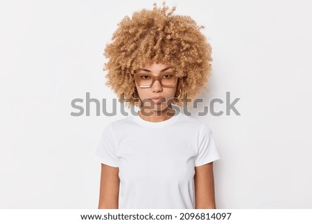Portrait of serious curly haired young woman looks attentively at camera wears spectacles and basic t shirt poses against white background has self confident expression listens you attentively