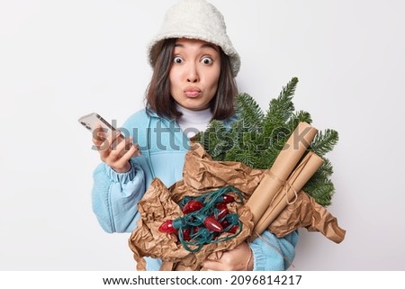 Photo of impressed lovely Asian woman stares bugged eyes has lips rounded reacts on something shocking prepares for coming holidays dressed in winter outerwear holds smartphone waits for call