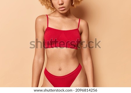Faceless fitness female model poses indoor. Unknown woman wears red top and panties has perfect body well cared smooth skin flat belly isolated over beige background demonstrates good shape.