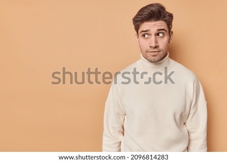 Horizontal shot of handsome dark haired man looks with curious expression at something interesting feels suspicious wears casual turtleneck isolated over beige wall with copy space area for text Royalty-Free Stock Photo #2096814283