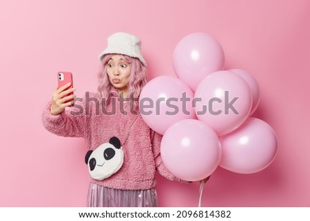 Lovely romantic Asian woman keeps lips rounded takes selfie via smartphone holds bunch of balloons going to congratulate friend with anniversary isolated over pink background. Celebration time
