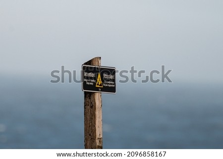 A sign posted which indicates "Attention Dangerous Coastline Stay on the designated trail" in both English and French text watching ocean waves. The background is blue ocean and grey fog. 