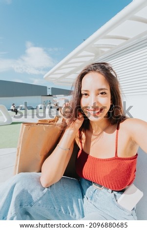 Curly hair african arab happy woman sitting outside shopping mall outdoors holding shopping bags take a selfie by camera, selfie concept, video call happy about his day. Urban look young people.