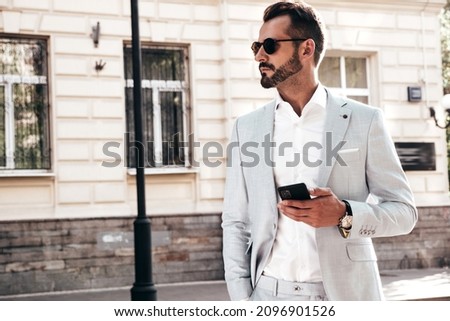 Portrait of handsome confident stylish hipster lambersexual model.Sexy modern man dressed in elegant suit. Fashion male posing in the street near fence at sunset. In sunglasses.Using smartphone apps Royalty-Free Stock Photo #2096901526