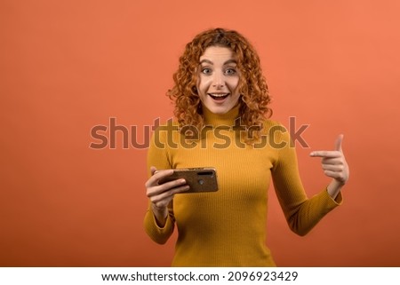 Young and attractive redhead Caucasian girl in orange jumper playing game on smartphone and pointing her finger at it isolated on orange studio background.