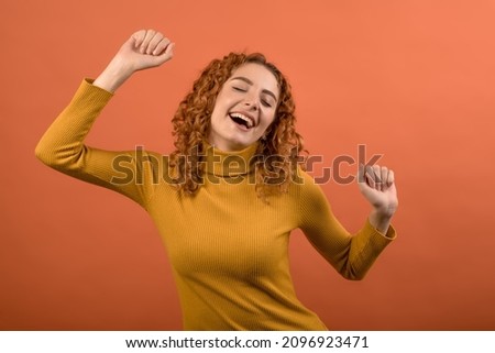 Attractive redhead caucasian young woman with curly hair in casual clothes with pleasure dancing and having fun at party isolated on orange studio background.