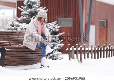 A girl laces up her skates while sitting on a park bench. A young woman came to skate at the rink.