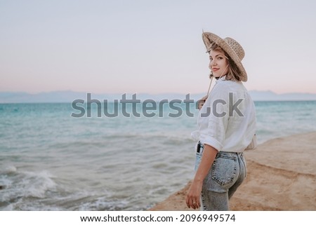 Pretty woman standing on beach near Mediterranean sea. Lady tourist in straw hat watching beautiful blue water surface, nature background. Windy weather, golden hour. High quality photo