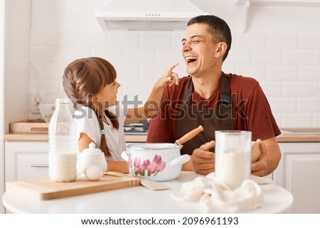 Laughing young adult dark haired man wearing maroon t shirt and apron cooking in kitchen with little daughter, kid smeared the dad's nose with flour, expressing happy emotions.