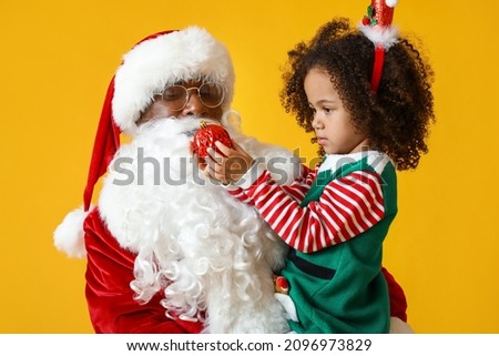 Little African-American girl with Santa Claus and Christmas ball on yellow background