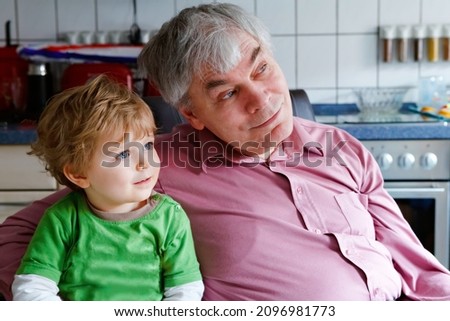 Little toddler boy and grandfather watching tv together. Happy family, grandchild and senior man, granddad at home, watch cartoons on television, indoors.