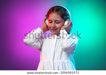 Half-length portrait of calm little girl, kid listening audio fairy-tail in headphones isolated on gradient purple blue background in neon. Human emotions, facial expression. Concept of childhood