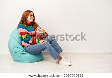 Young caucasian woman sitting on puff with popcorns isolated on white background looks aside smiling, cheerful and pleasant.
