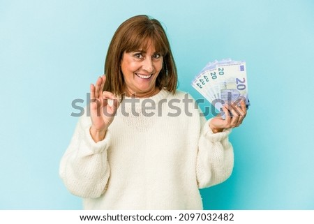 Middle age caucasian woman holding bank notes isolated on blue background cheerful and confident showing ok gesture.