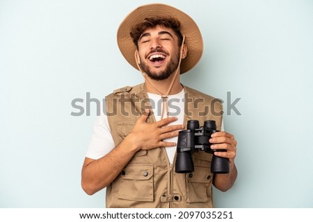 Young mixed race man holding binoculars isolated on blue background laughs out loudly keeping hand on chest.
