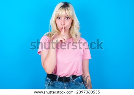 Caucasian girl wearing pink T-shirt isolated over blue background  makes hush gesture, asks be quiet. Don't tell my secret or not speak too loud, please!
