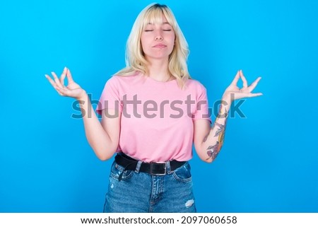 Caucasian girl wearing pink T-shirt isolated over blue background  doing yoga, keeping eyes closed, holding fingers in mudra gesture. Meditation, religion and spiritual practices.