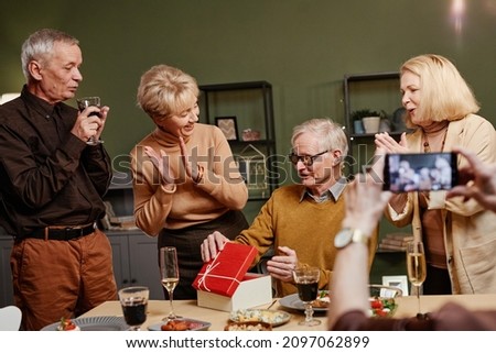 Joyful senior Caucasian man opening present box while sitting at table during birthday celebration with friends. Unrecognizable woman shooting using smartphone camera