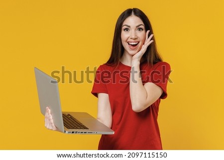 Shocked surprised amazed astinished charming young brunette woman 20s wears basic red t-shirt hold use work on laptop pc computer touch cheek with hand isolated on yellow background studio portrait