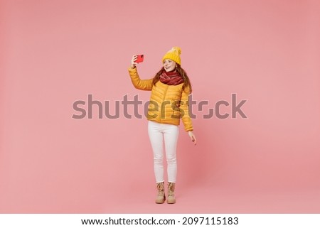 Full size body length young woman 20s wears yellow jacket hat mittens doing selfie shot on mobile cell phone post photo on social network isolated on plain pastel light pink background studio portrait