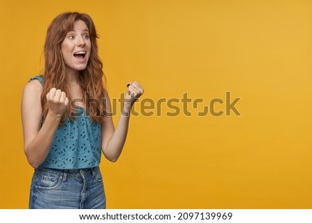 Indoor portrait of young redhead female with wavy long hair shouting and shows victory sign while watching football. isolated over orange background