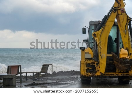 Close-up of a yellow tractor clearing the embankment of sand and mud after a storm against the background of the still raging sea, copy space