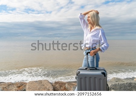 Close up young blonde woman in jeans and a white shirt with a raised hand on her head with a suitcase stands on huge rocks near the sea in cloudy weather and looks away