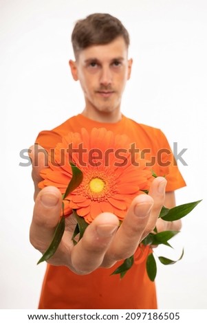 Young handsome tall slim white man with brown hair holding orange flower in his hand with orange shirt isolated on white background