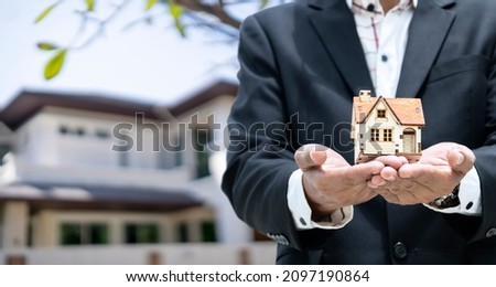 Businesses on buying - selling homes, businessmen with models, house put on hand, backdrop being home, wearing black suit, white shirt