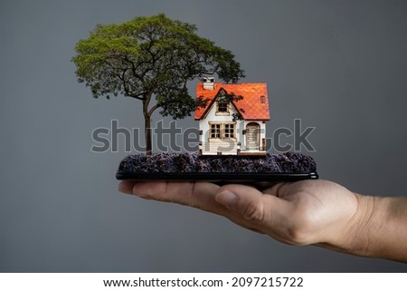 The businessman's hand holds a smartphone and then there is land, trees, houses and small apartments on it. Smartphone application for online searching, buying, selling and booking real estate