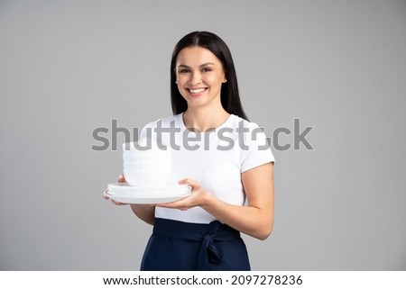 Waist up portrait view of the attractive woman waitress holding a lot of empty plates and smiling to the camera isolated on grey background. Stock photo 